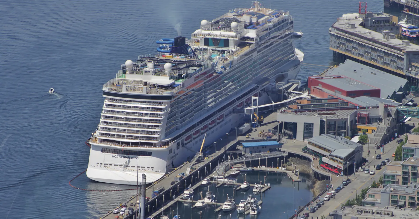 A large cruise ship tied up at Pier 66, the Bell Street cruise terminal on Seattle's downtown waterfront, seen from above, with a small marina and the terminal buildings beside it