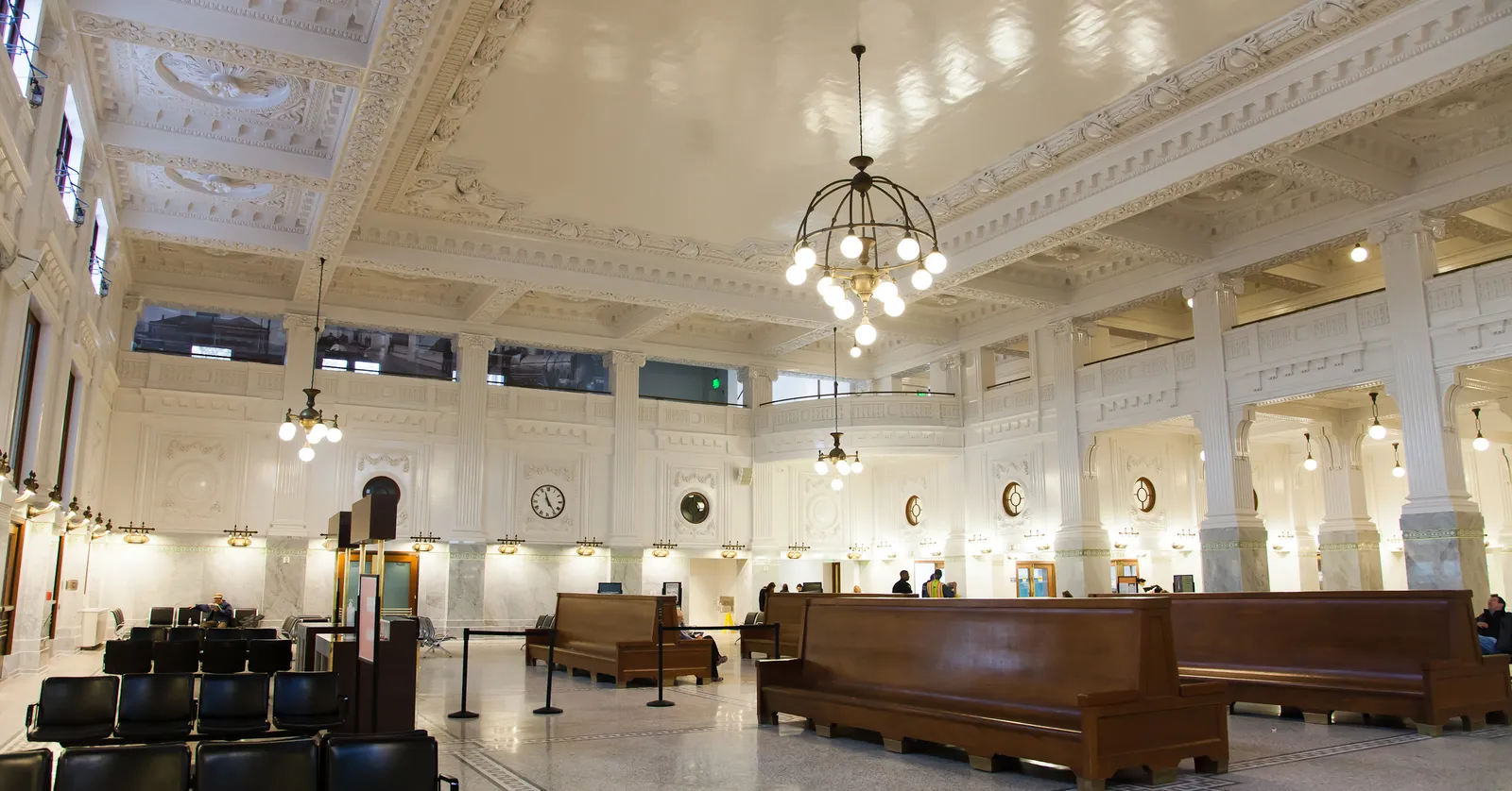 The restored waiting room at Seattle's King Street Station, with an ornamental white plaster ceiling, a bronze chandelier, wooden benches and a terrazzo floor