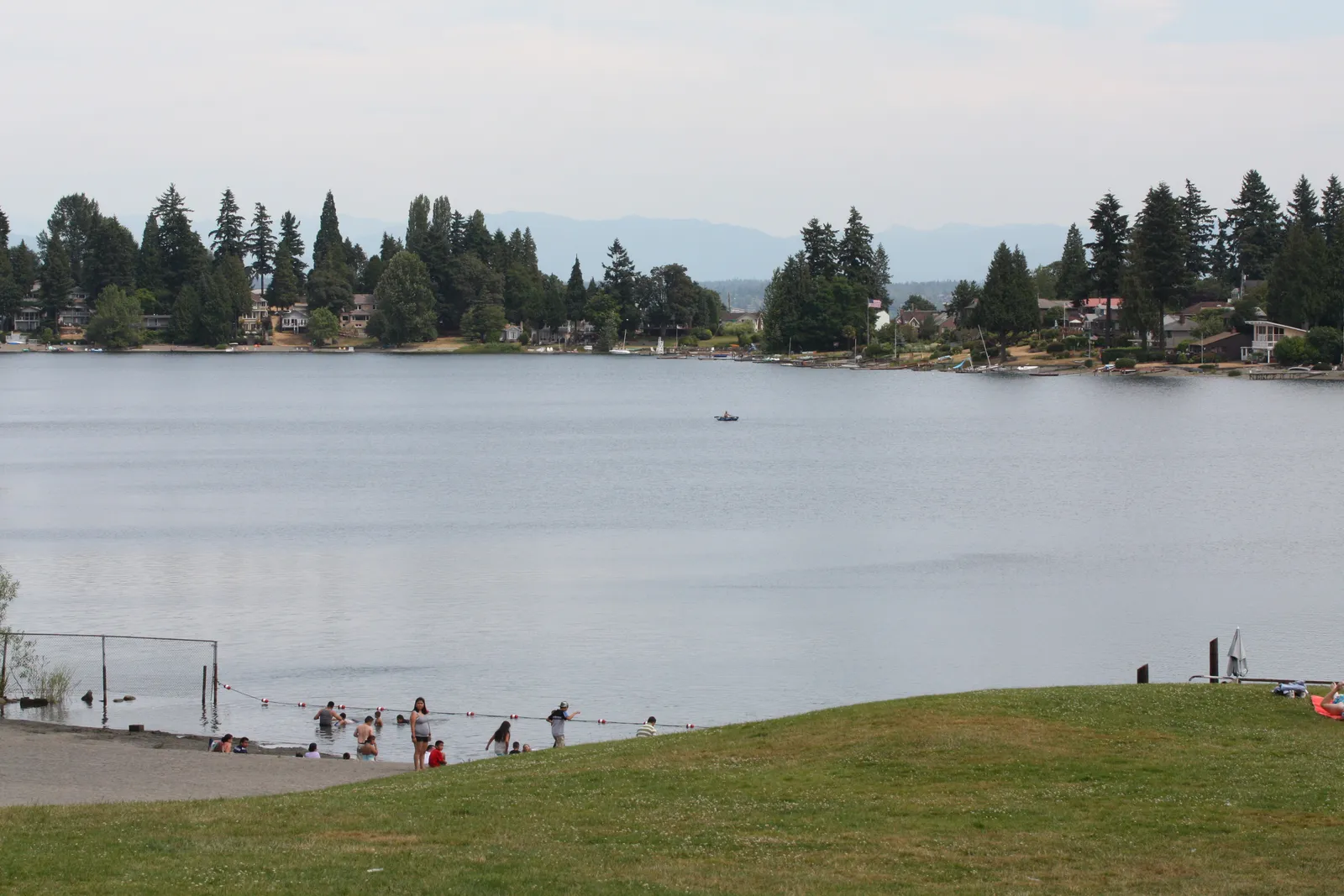 Angle Lake in SeaTac, Washington, with a roped swimming area, a grass bank in the foreground and wooded shoreline houses across the water