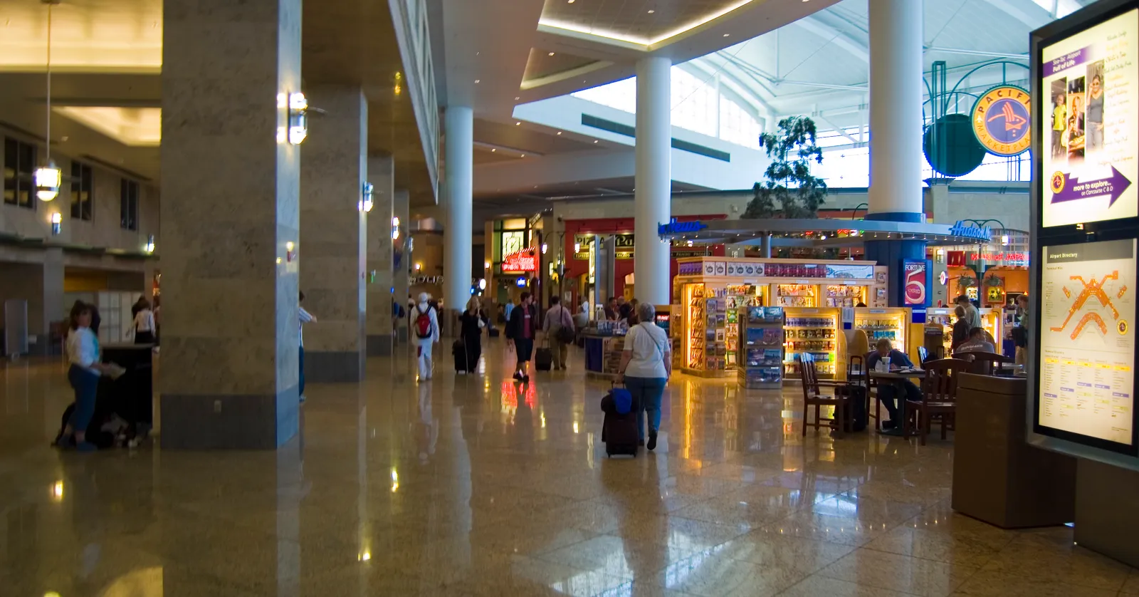 A concourse inside Seattle-Tacoma International Airport, with shops along one side, a polished stone floor and travelers wheeling bags past