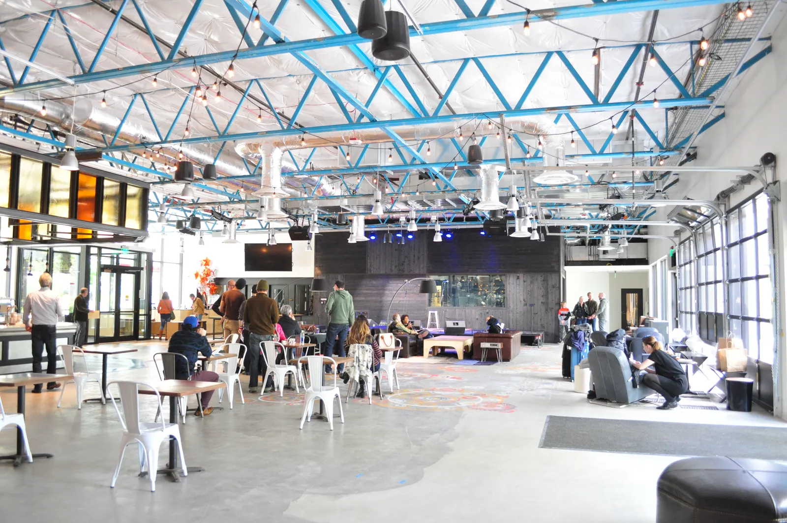 KEXP's public gathering space at Seattle Center, a high open room with blue roof trusses, cafe tables and a small stage at the far end