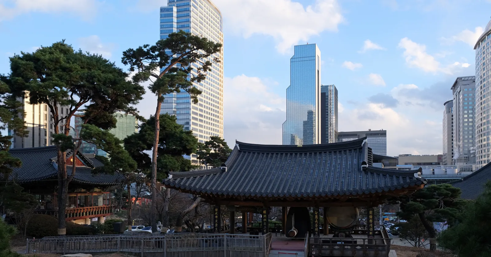 A tiled-roof hall at Bongeunsa temple in Seoul among pine trees, with the glass office towers of Gangnam rising directly behind it