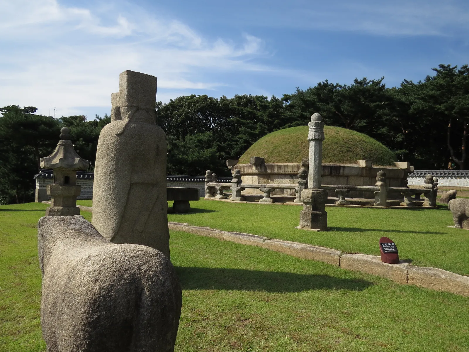 A stone horse and a carved stone official standing on the lawn in front of a grassy royal burial mound at the Seonjeongneung tombs in Seoul, with pines behind