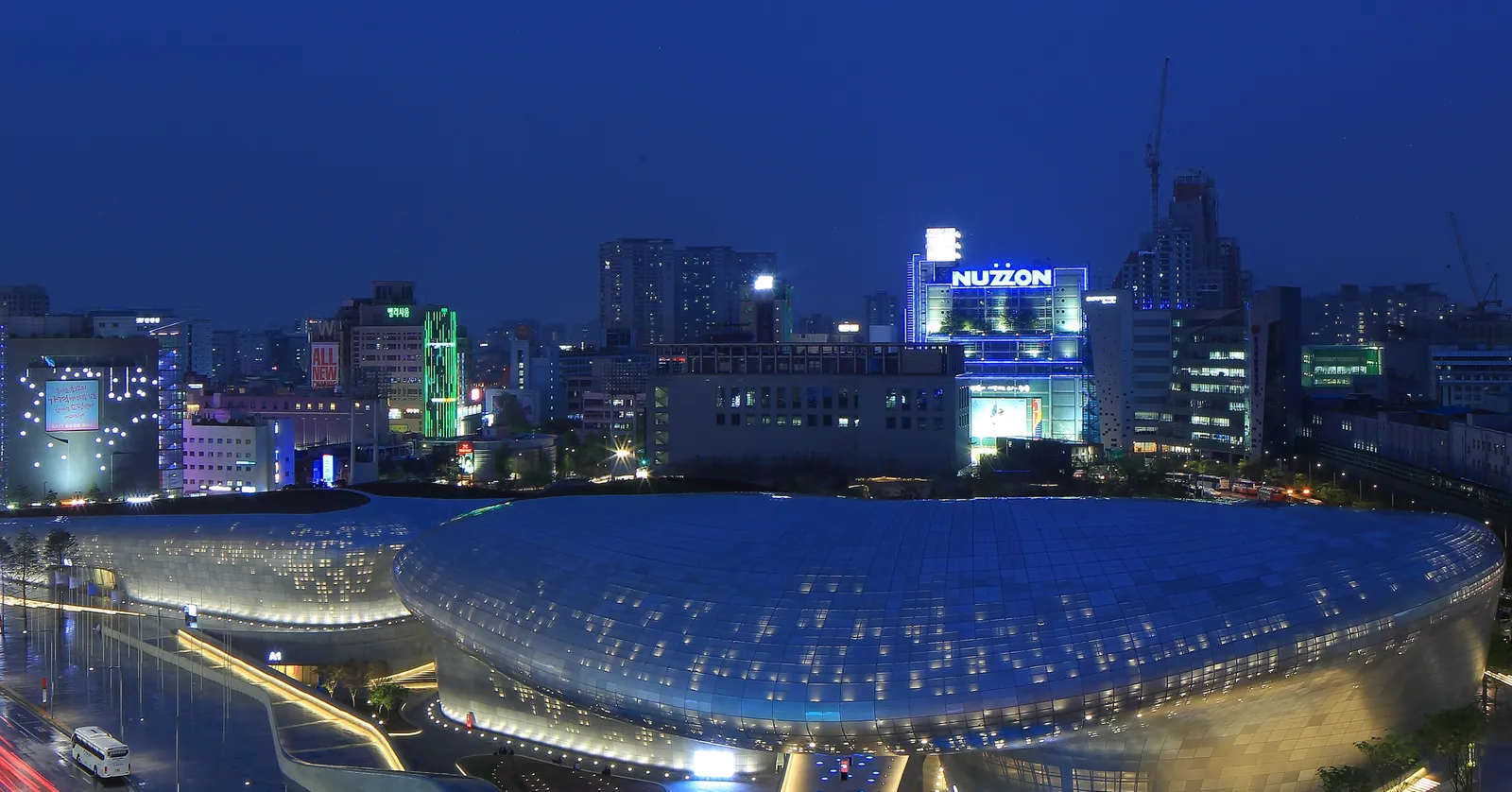 Dongdaemun Design Plaza in Seoul at dusk, its long, curving metal-panelled roof lit from within, with neon-lit shopping towers behind it