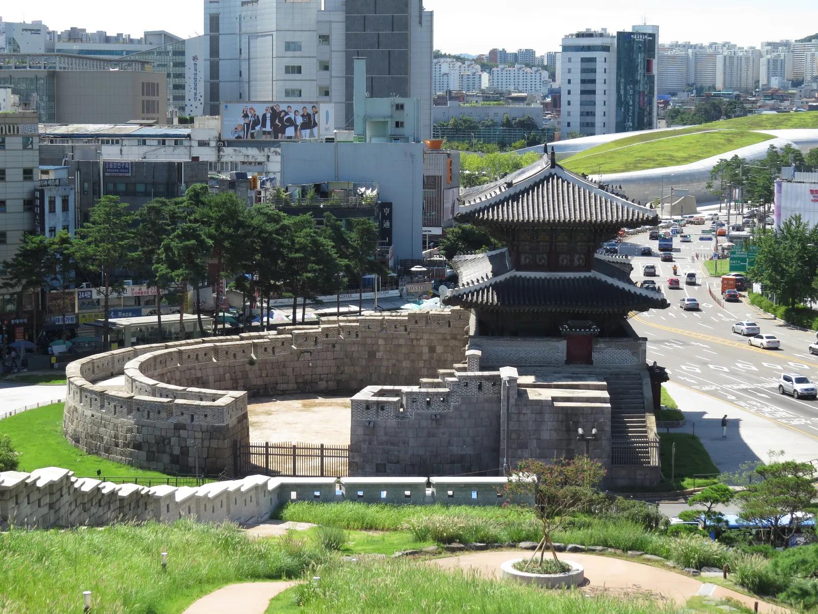 Heunginjimun, Seoul's east gate, a two-storey wooden gatehouse on a stone base wrapped by a curved half-moon outer wall, with traffic passing and buildings behind