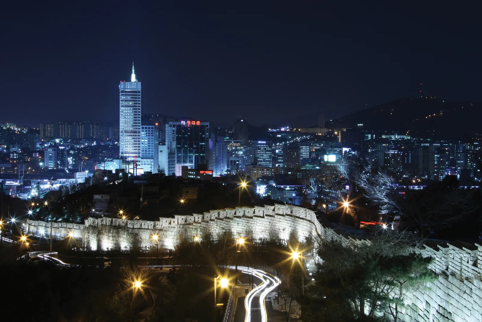 The Seoul City Wall on Naksan at night, a floodlit line of stone battlements curving along a dark hillside, with lit towers of the city behind