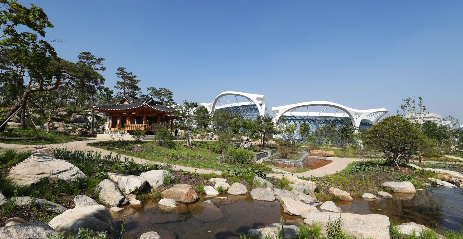 The outdoor gardens at Seoul Botanic Park, a rock-edged pond and pine trees in front of a traditional Korean pavilion, with the white curved roofs of the greenhouse beyond