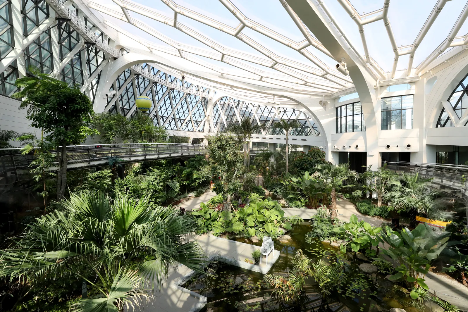 Inside the greenhouse at Seoul Botanic Park, palms and tropical plants under a white lattice roof of glass panels, with a raised walkway running around the upper level