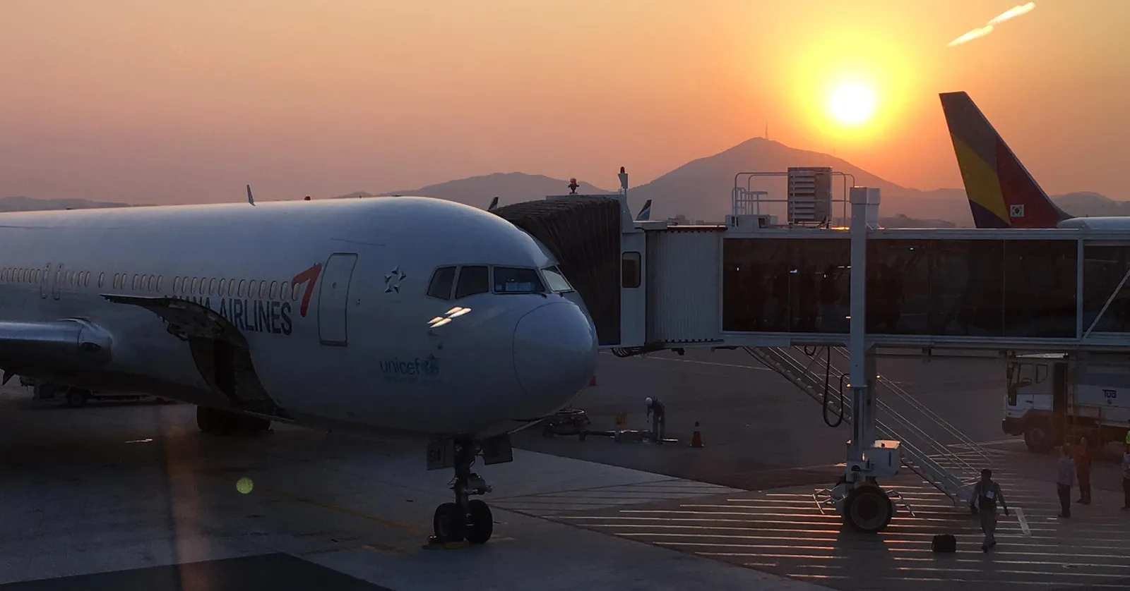 An airliner parked at a jet bridge at Gimpo International Airport in Seoul at sunset, with a hill on the horizon behind the terminal and the sun low in an orange sky