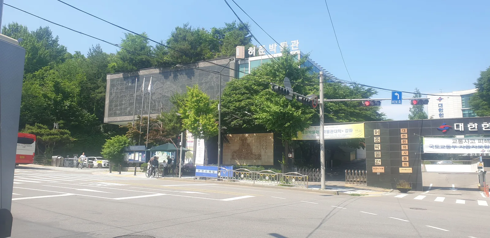The front of the Heojun Museum in Gangseo, Seoul, a low modern building behind a gated entrance and trees, seen from across a street crossing