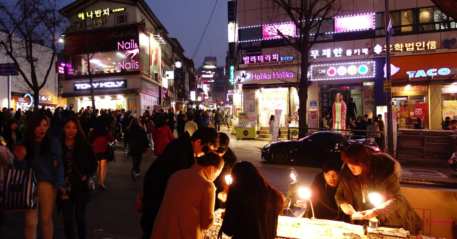 A Hongdae street in Seoul at night, lit shopfronts and neon signs above a crowd, with street vendors selling under lamps on the pavement in the foreground