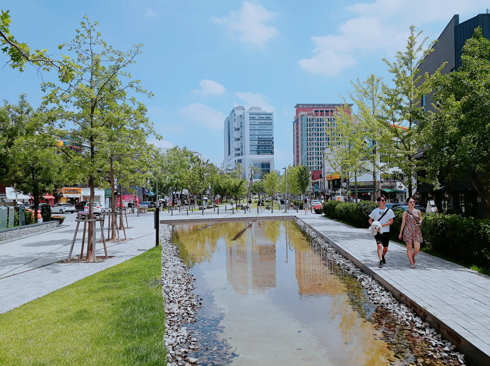 The Yeonnam-dong section of Gyeongui Line Forest Park in Seoul, a long shallow stream between paved paths, lawns and young trees, with office towers at the far end