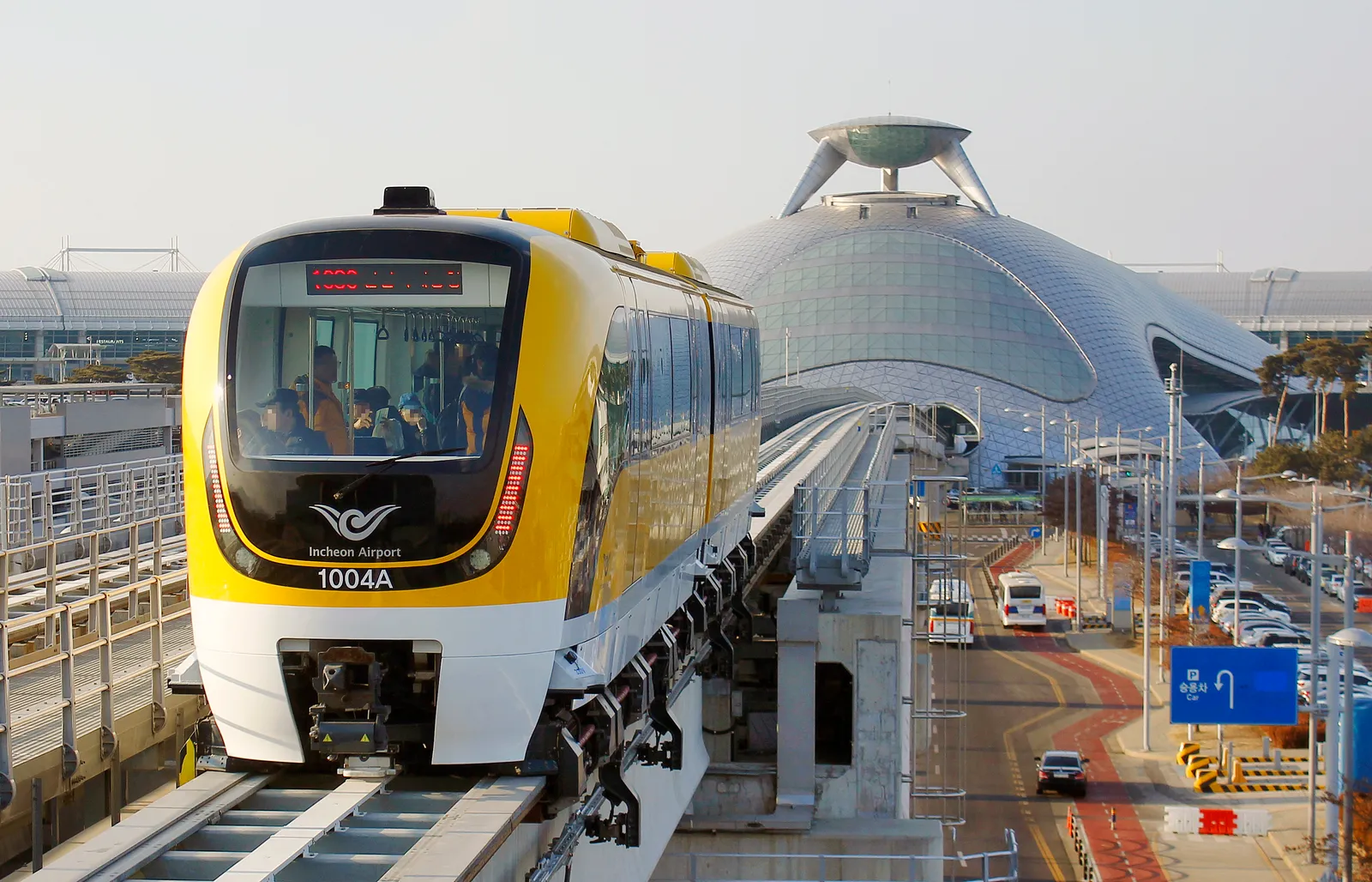 A yellow and white two-car maglev train on an elevated guideway outside Incheon Airport Terminal 1, with the terminal's curved roof behind it