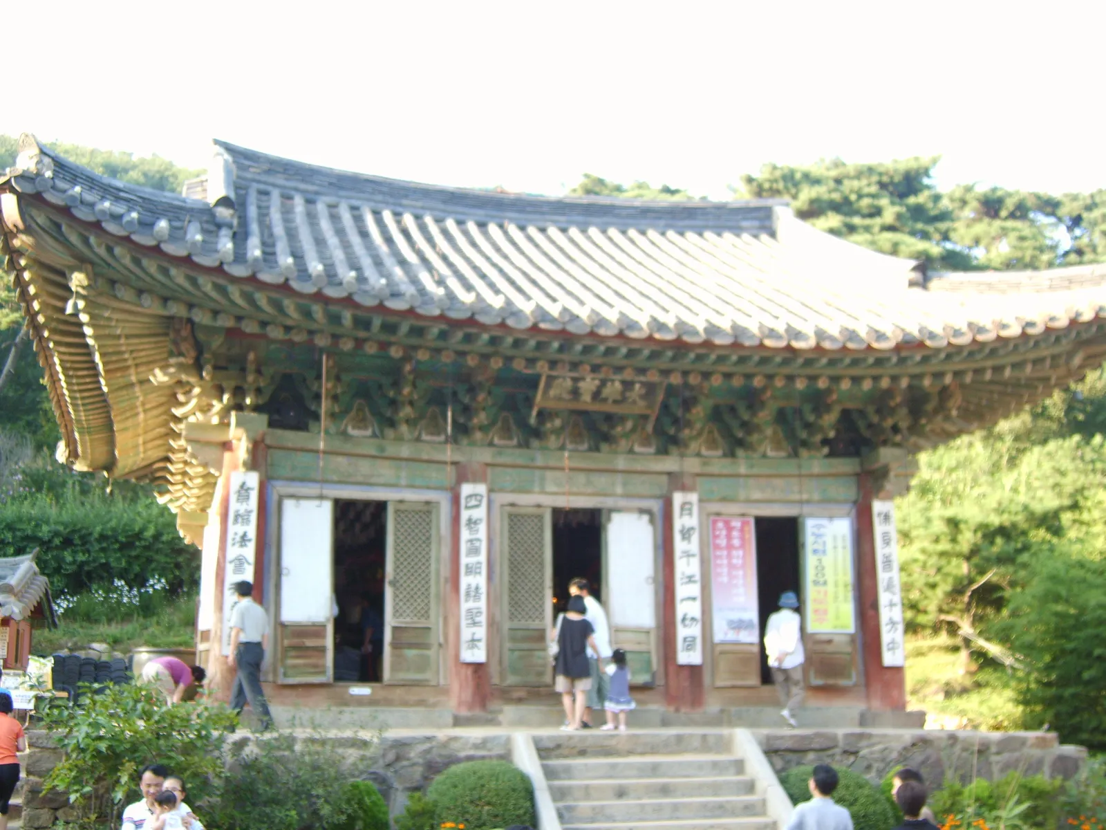A small wooden hall at Jeondeungsa temple on Ganghwa Island, with painted brackets under a tiled roof and visitors climbing the stone steps to its doors among trees