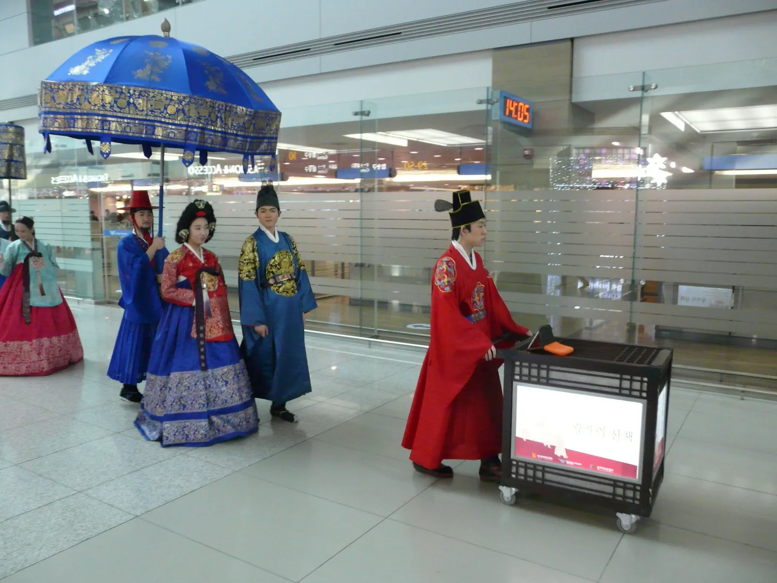 Performers in Joseon-era royal costume, a king and queen under a blue ceremonial parasol with attendants in red and blue robes, walking through the departure hall at Incheon Airport