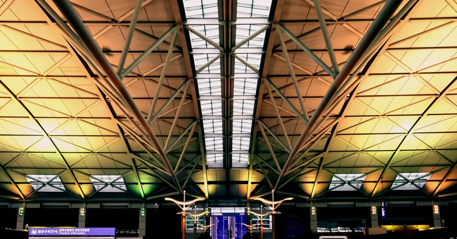 The steel-trussed roof of the check-in hall at Incheon International Airport Terminal 1, lit gold and green, with a long skylight down the middle and departure boards below