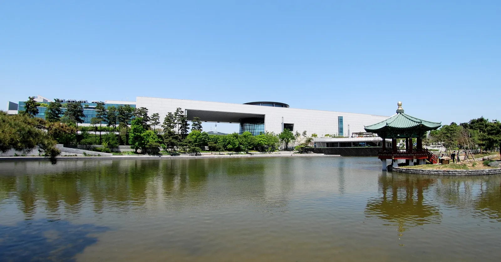 The long pale stone building of the National Museum of Korea in Seoul seen across Mirror Pond, with a small pavilion on the water to the right and trees along the bank