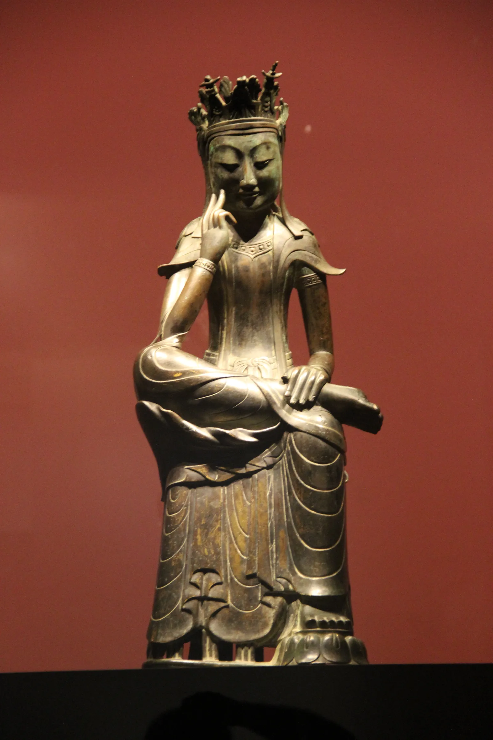 A gilt-bronze pensive bodhisattva of the late sixth century at the National Museum of Korea, seated with one leg crossed over the other and fingers raised to the cheek, against a dark red wall
