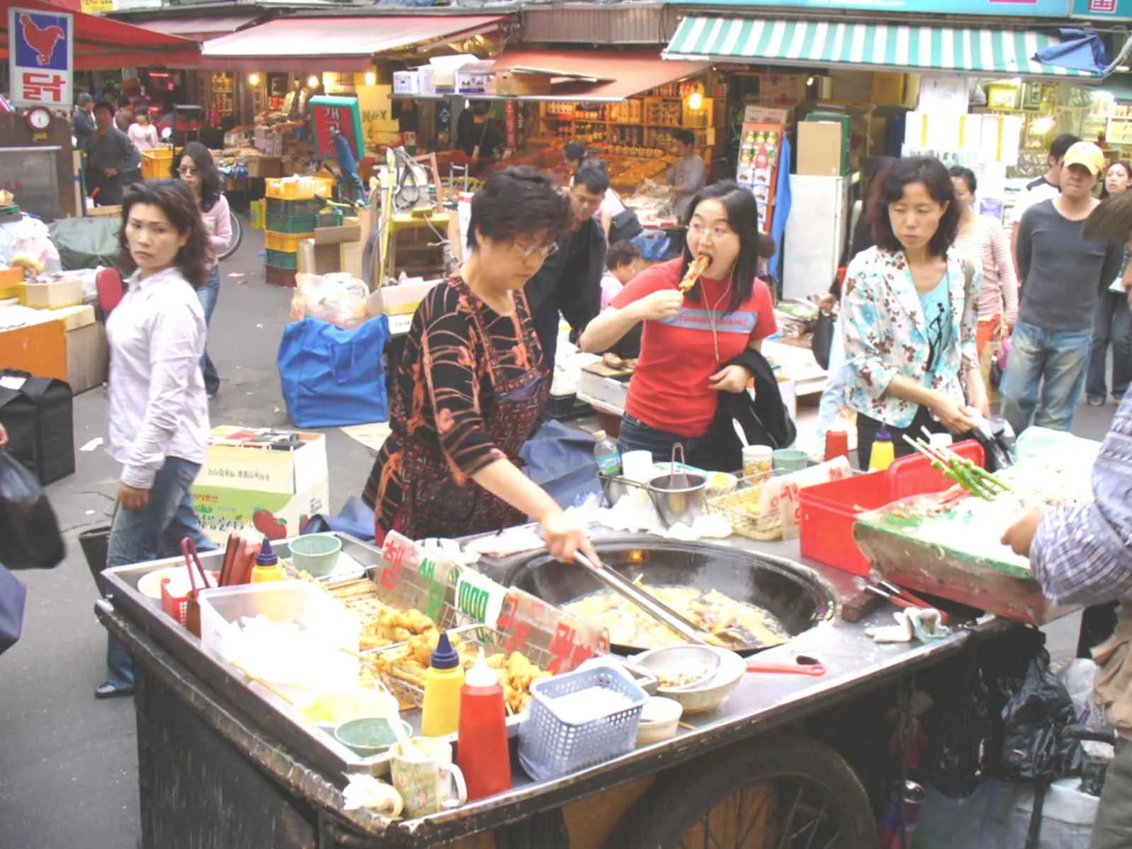 Street food carts in Namdaemun Market in Seoul, a vendor cooking at a wheeled stall under striped awnings with shoppers crowding the lane around it