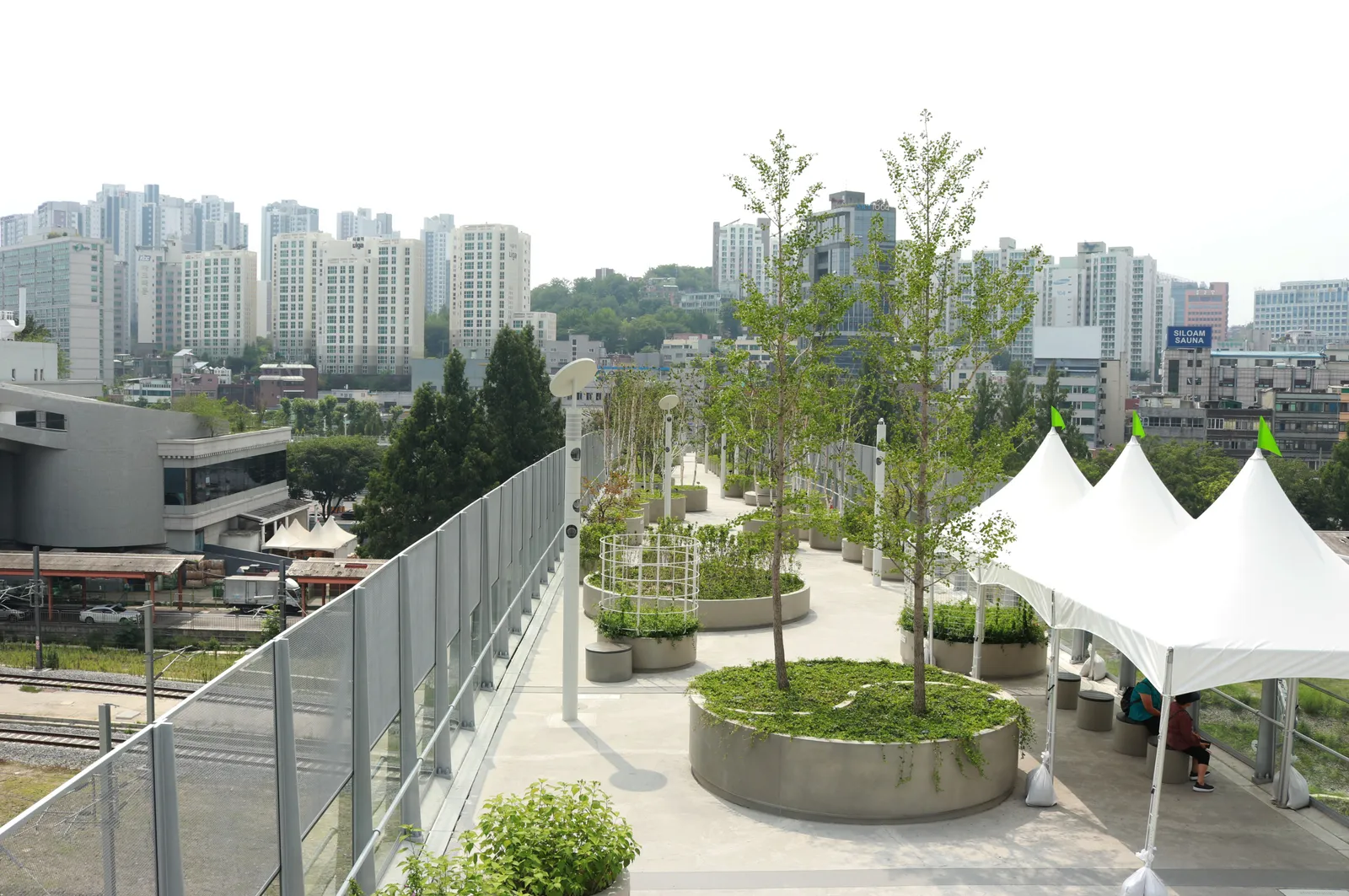 The Seoullo 7017 elevated walkway in Seoul, a pale concrete deck with round planters holding young trees and shrubs, running beside railway tracks with apartment towers behind