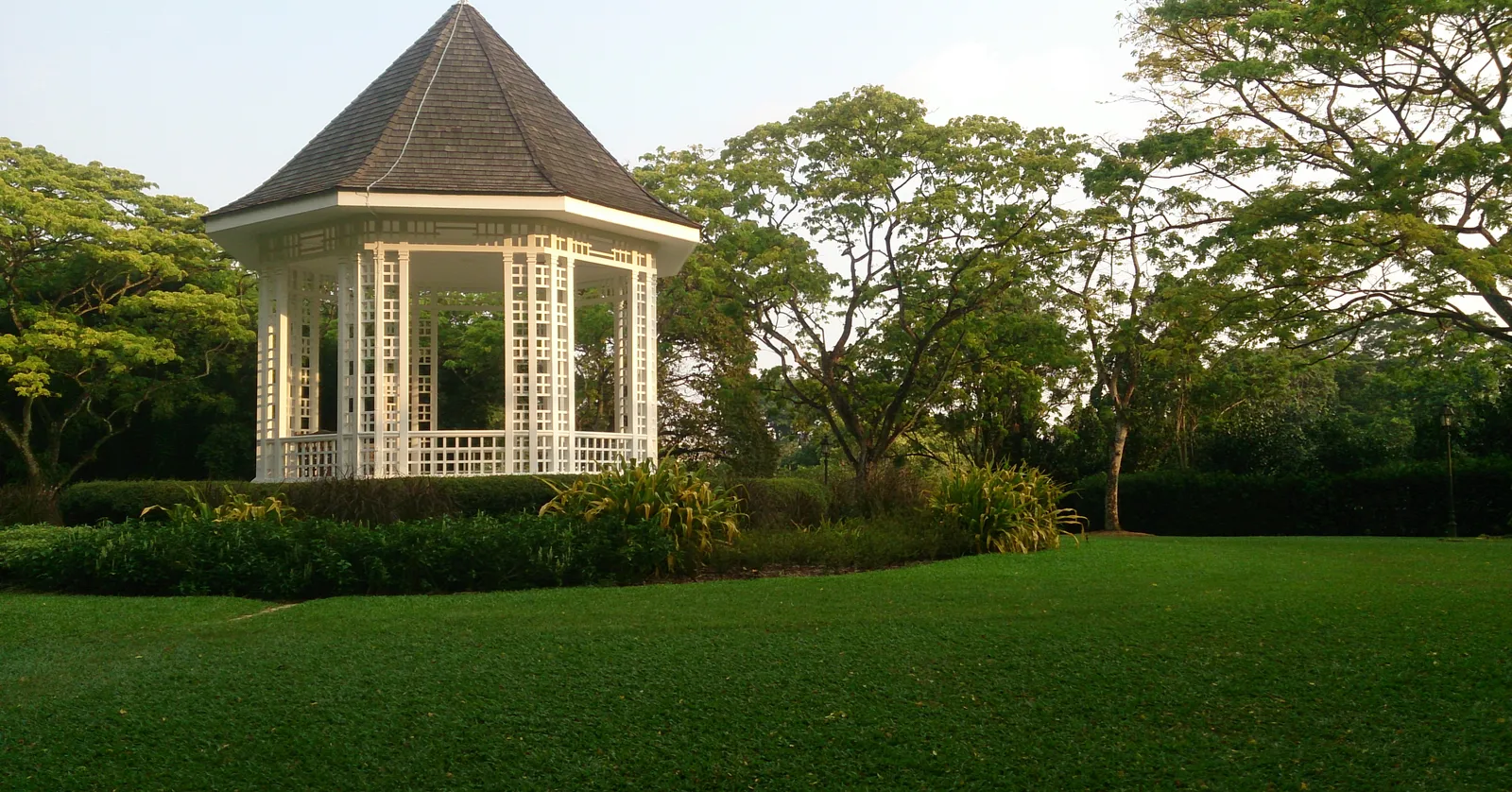 The white latticework bandstand at Singapore Botanic Gardens standing on a mown lawn under a conical roof, mature trees closing in behind it