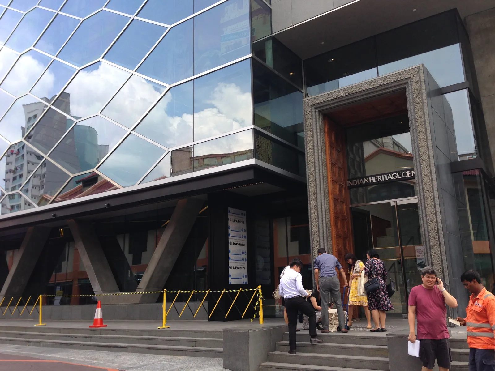 The Indian Heritage Centre on Campbell Lane in Singapore, a faceted glass and steel facade angled over the pavement beside a dark stone entrance, with visitors on the steps