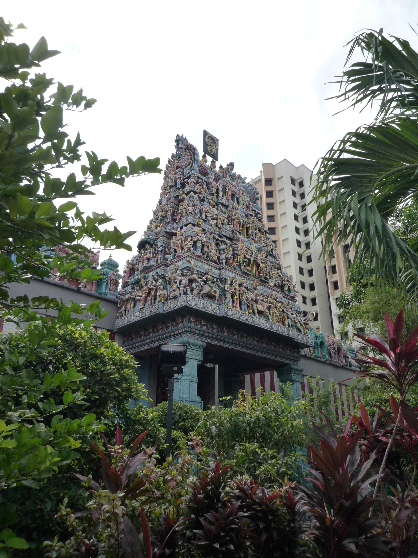 Sri Veeramakaliamman Temple on Serangoon Road in Singapore, a stepped gopuram tower covered in painted figures rising over the temple entrance, framed by palms with a housing block behind
