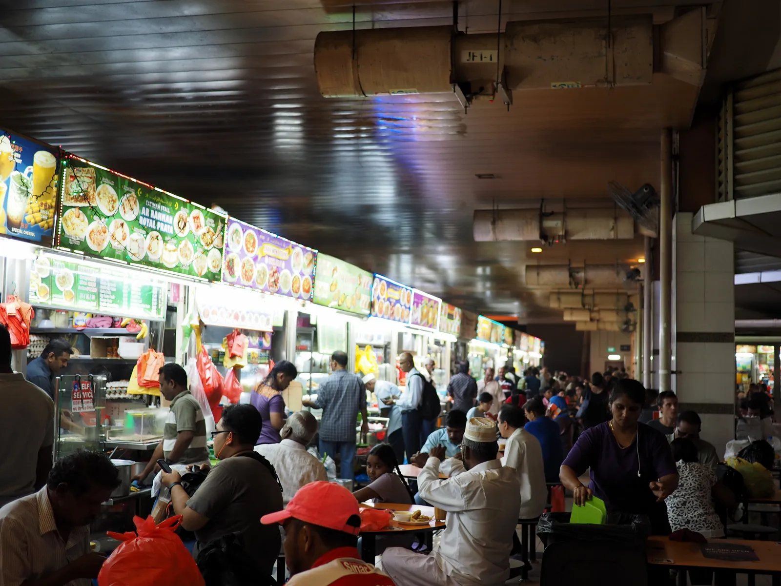 The hawker section inside Tekka Centre in Singapore, lit stall signboards above steel counters down one side and crowded tables of diners along the other