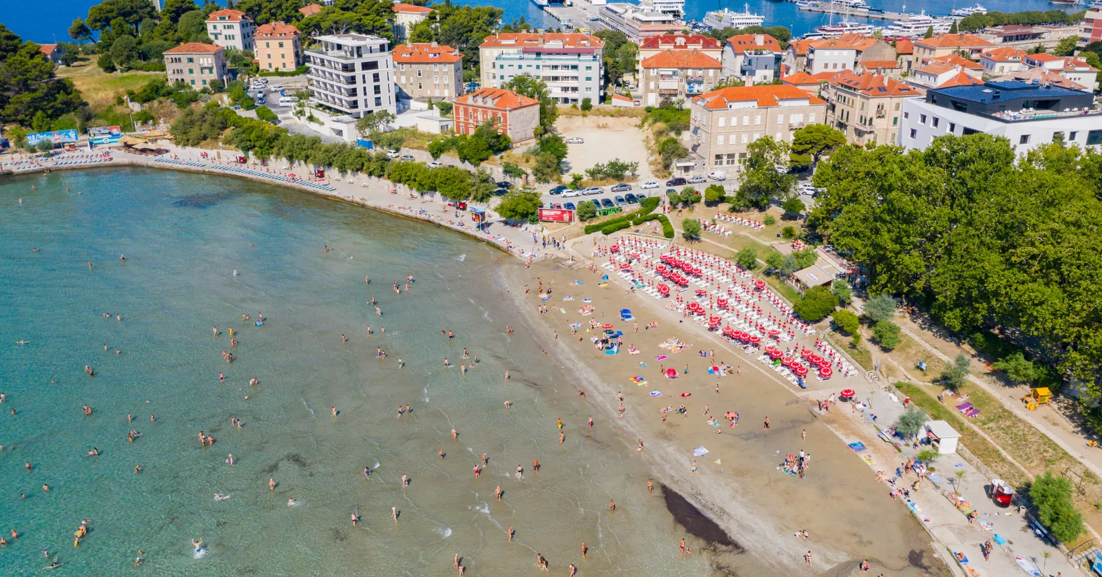 Bačvice beach in Split from above, a broad crescent of pale sand crowded with sunbathers and rows of red parasols, shallow turquoise water full of swimmers, and the roofs of the city behind