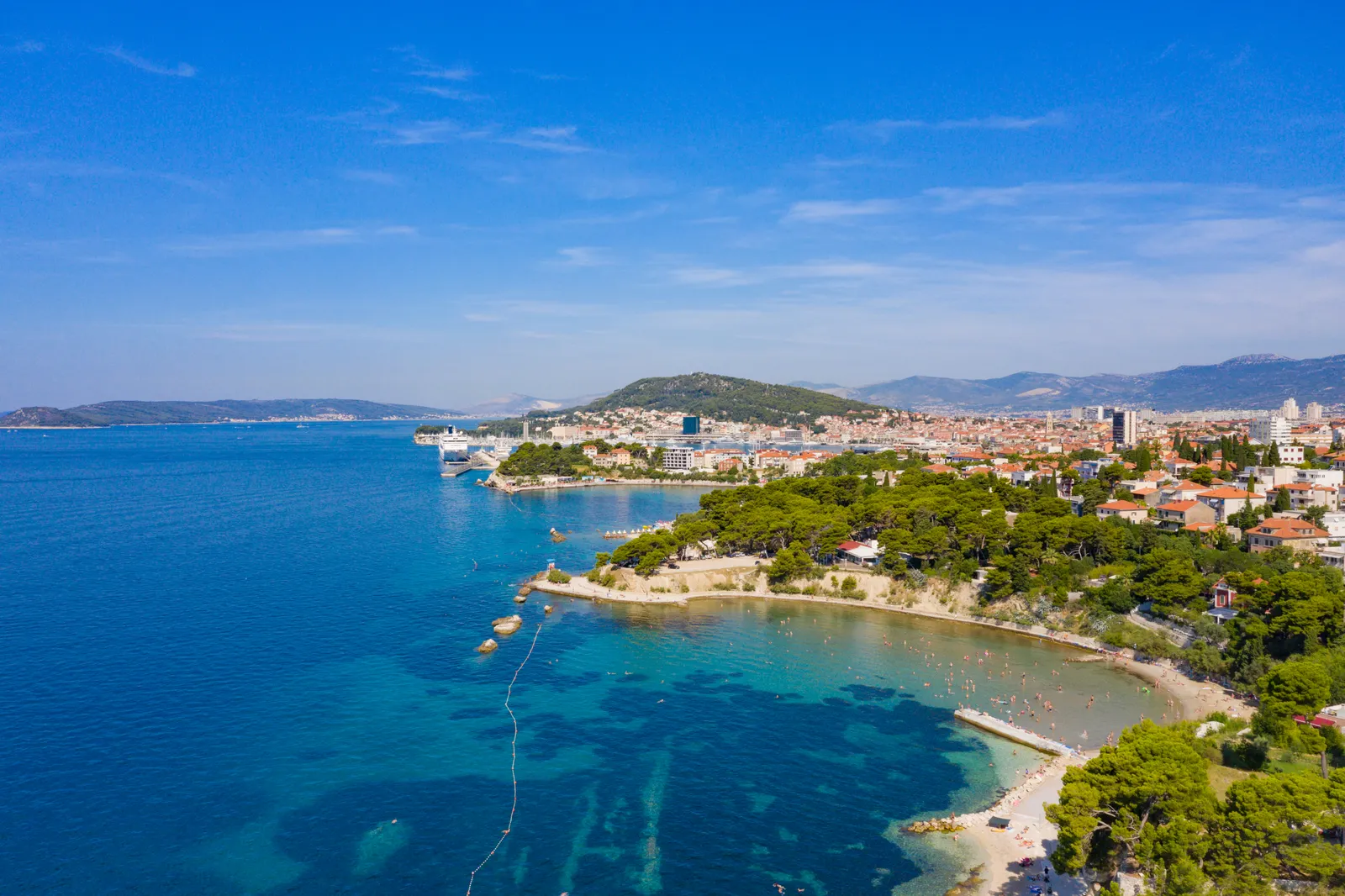 A pine-covered headland on Split's eastern shore with a small sheltered cove and clear turquoise water, the city's apartment blocks and harbour spread out behind it under a blue sky