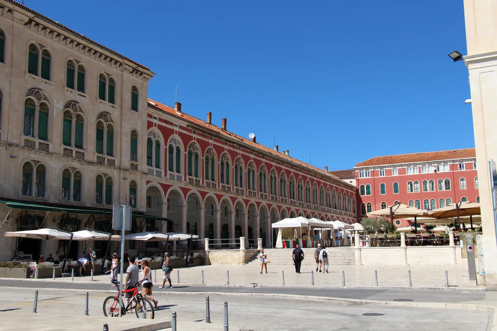 Prokurative square in Split, a long red building with two tiers of green-shuttered arches down one side and a pale stone palace on the other, café awnings under them and people crossing the open paving