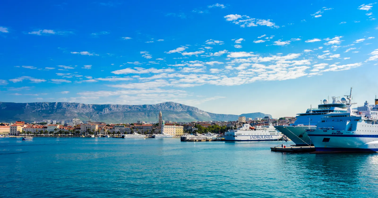 Split's harbour from the water, a white Jadrolinija ferry and a larger cruise ferry moored along the quay on the right, the low ochre and cream buildings of the old town behind them and a long grey mountain ridge beyond