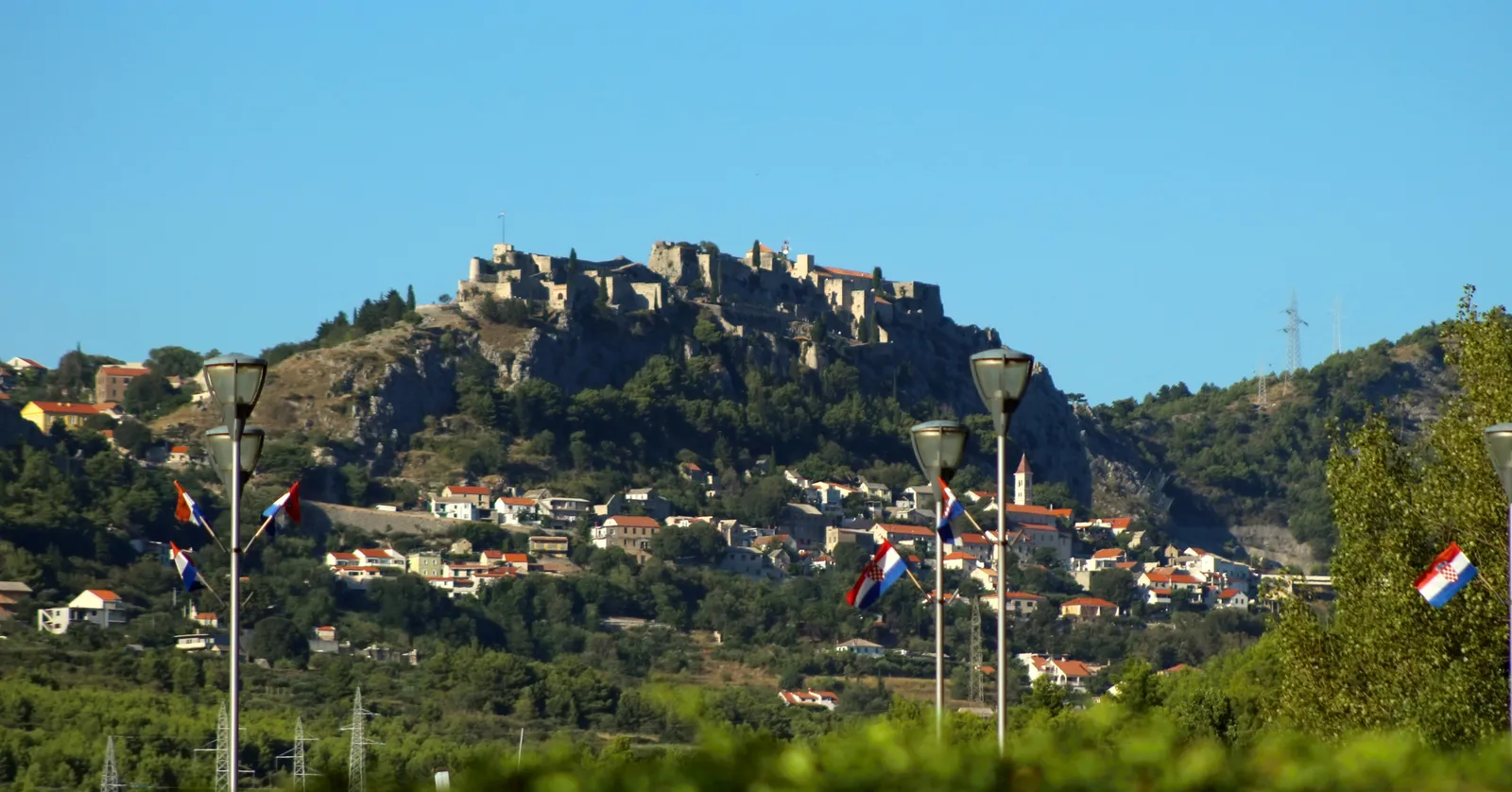 Klis Fortress seen from below, a long grey stone fortress running along the crest of a rocky ridge above a village of red-roofed houses, with Croatian flags on poles in the foreground