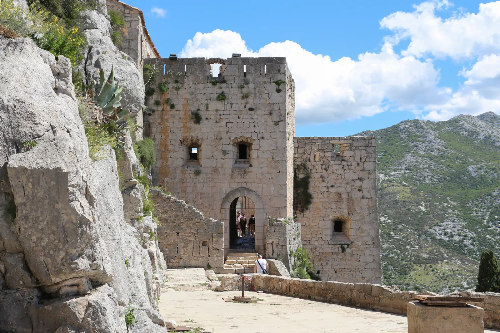 A square stone gate tower inside Klis Fortress with an arched doorway through it, weathered rampart walls and an agave growing out of the rock on the left, bare limestone mountains beyond