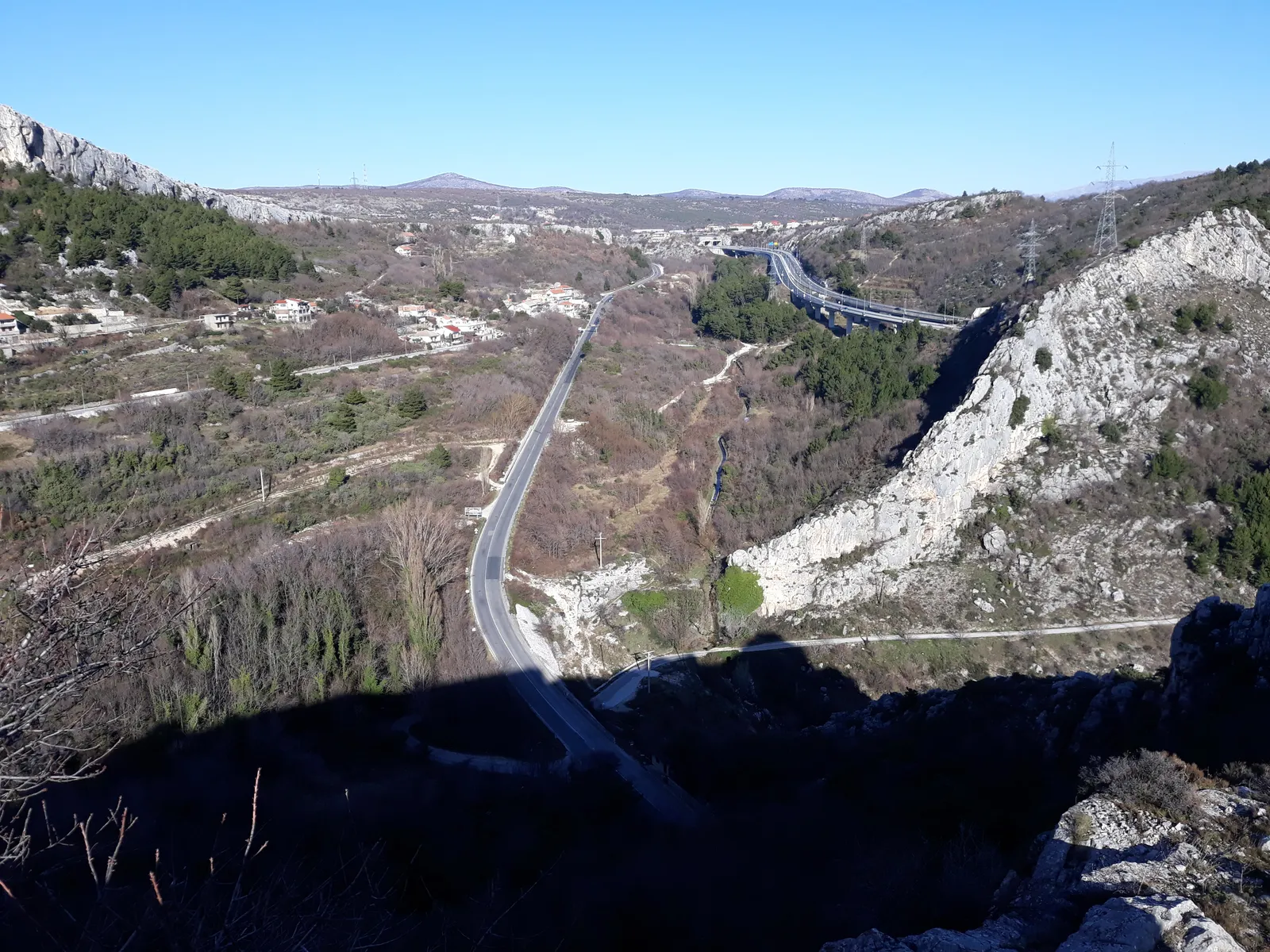 The view north from Klis over the pass, a road winding down between bare grey karst ridges into a wide dry valley with scattered houses and a motorway viaduct in the distance