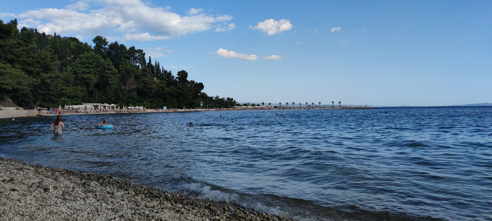 Kašjuni cove below Marjan in Split, a curve of white pebbles running into clear water with dense pine woods coming down to the shoreline and the open sea beyond