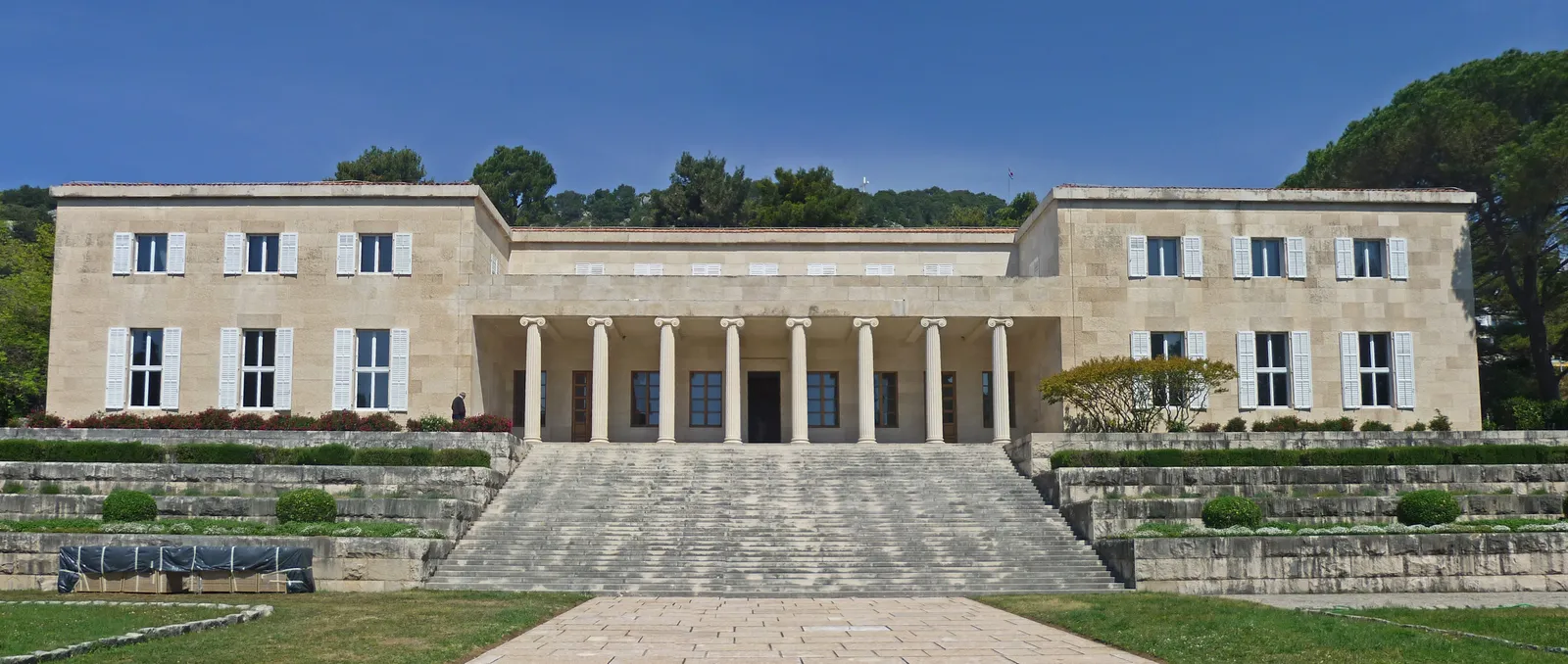 The Meštrović Gallery in Split, a long symmetrical pale stone house with a colonnaded portico at its centre, standing above wide terraced steps and clipped garden beds with pine woods behind