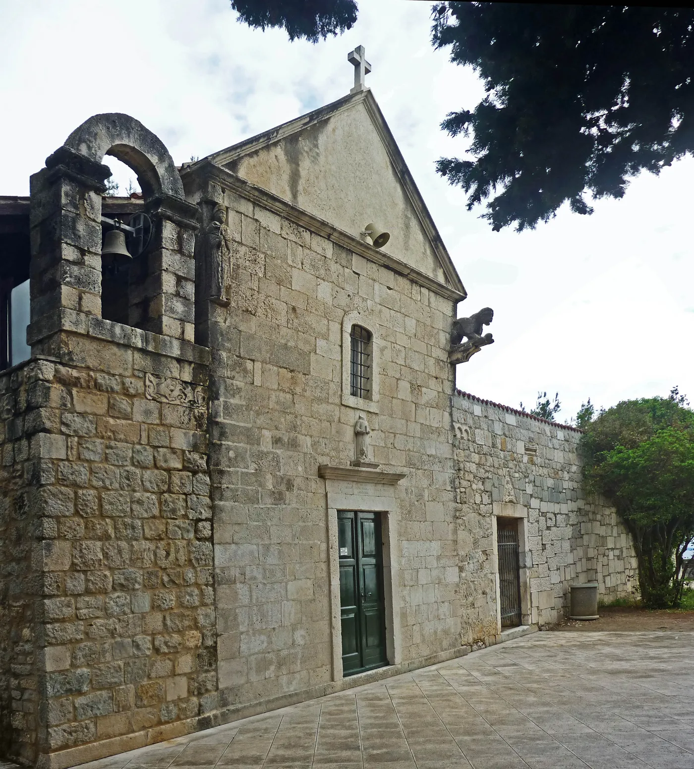 A small stone church on the Sustipan headland in Split, plain gabled front with a single green door and an open arch holding a bell beside it, cypress and pine trees overhanging the paved terrace