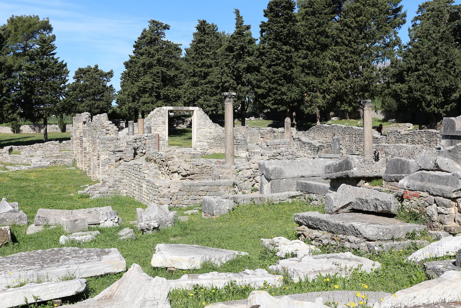The ruins of the cemetery basilica at Manastirine in Salona, low stone walls and two standing columns among dark cypress trees, with carved stone sarcophagi lying in the grass in the foreground