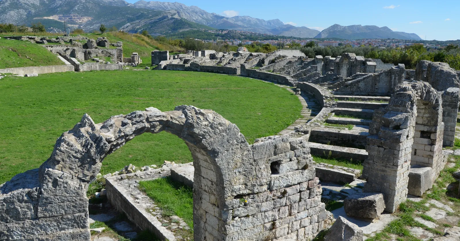 The ruined Roman amphitheatre at Salona, a grass oval ringed by broken limestone arches and tiers of seating stubs, with the bare grey mountains behind Solin rising in the distance