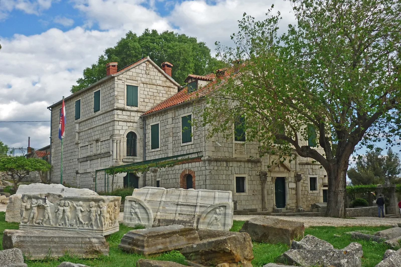 The Tusculum house at Salona, a two-storey stone building with green shutters and a red-tiled roof, with Roman sarcophagi carved with figures standing on the grass in front of it