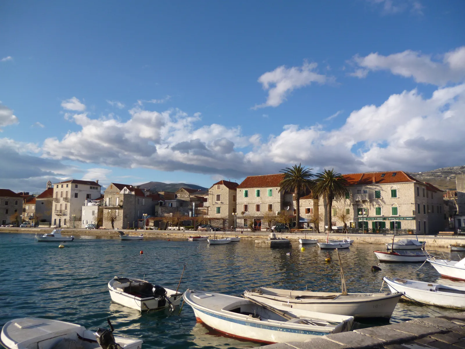 The waterfront at Kaštel Stari, small open boats moored in the shallow harbour in front of a row of stone houses with terracotta roofs, a palm tree among them and hills behind