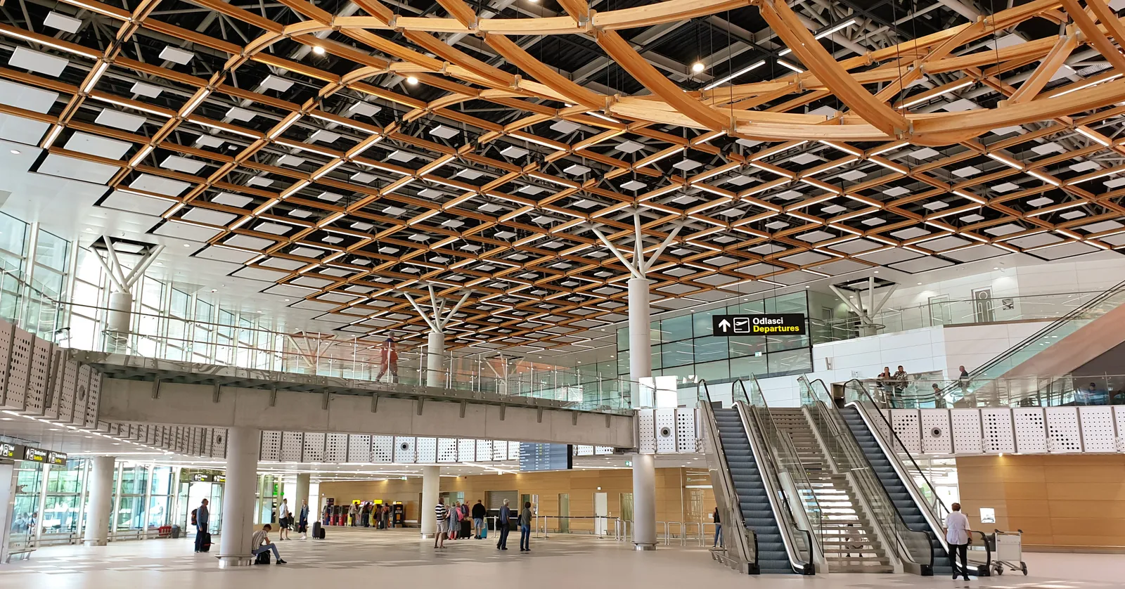 The main hall of Split Airport's terminal, a wide concourse under a lattice ceiling of pale timber beams, with escalators up to the departures level on the right and passengers crossing the floor below