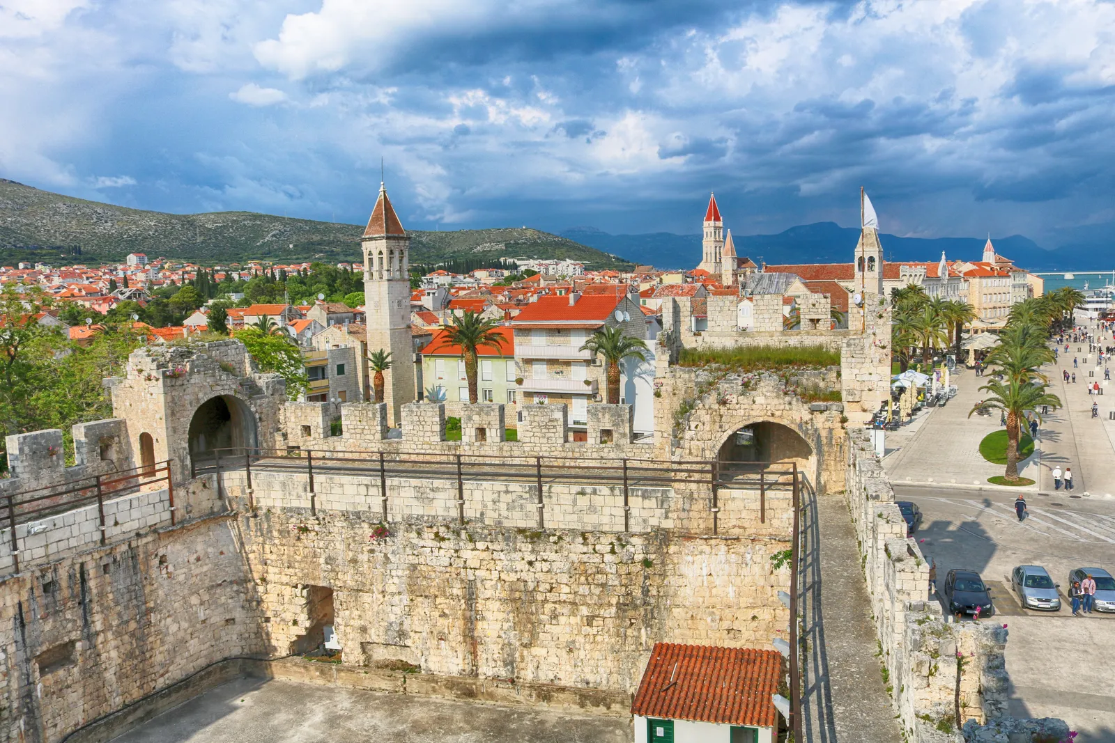 The view from the walls of Kamerlengo fortress in Trogir, broad stone ramparts in the foreground and the town's terracotta roofs, bell towers and palm-lined waterfront spread out beyond under a stormy sky