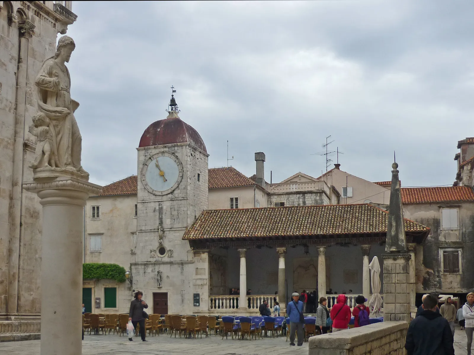 Trogir's main square, an open-sided stone hall with a tiled roof carried on plain columns, a clock tower with a red dome beside it, café tables on the paving and a statue on a column in the foreground
