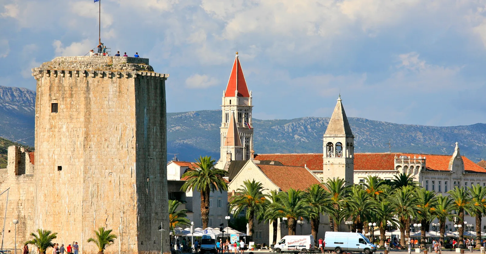 Trogir's waterfront, a square battlemented stone tower on the left with people on its roof, the cathedral's tall bell tower with a red pyramid roof behind the palm-lined promenade, and grey hills across the water