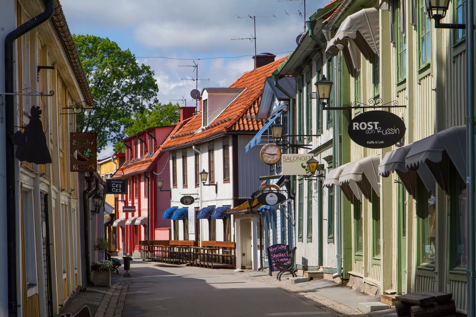 Stora gatan in Sigtuna, a narrow street of low wooden houses painted yellow, red and green with tiled roofs and wrought-iron shop signs hanging out over the pavement