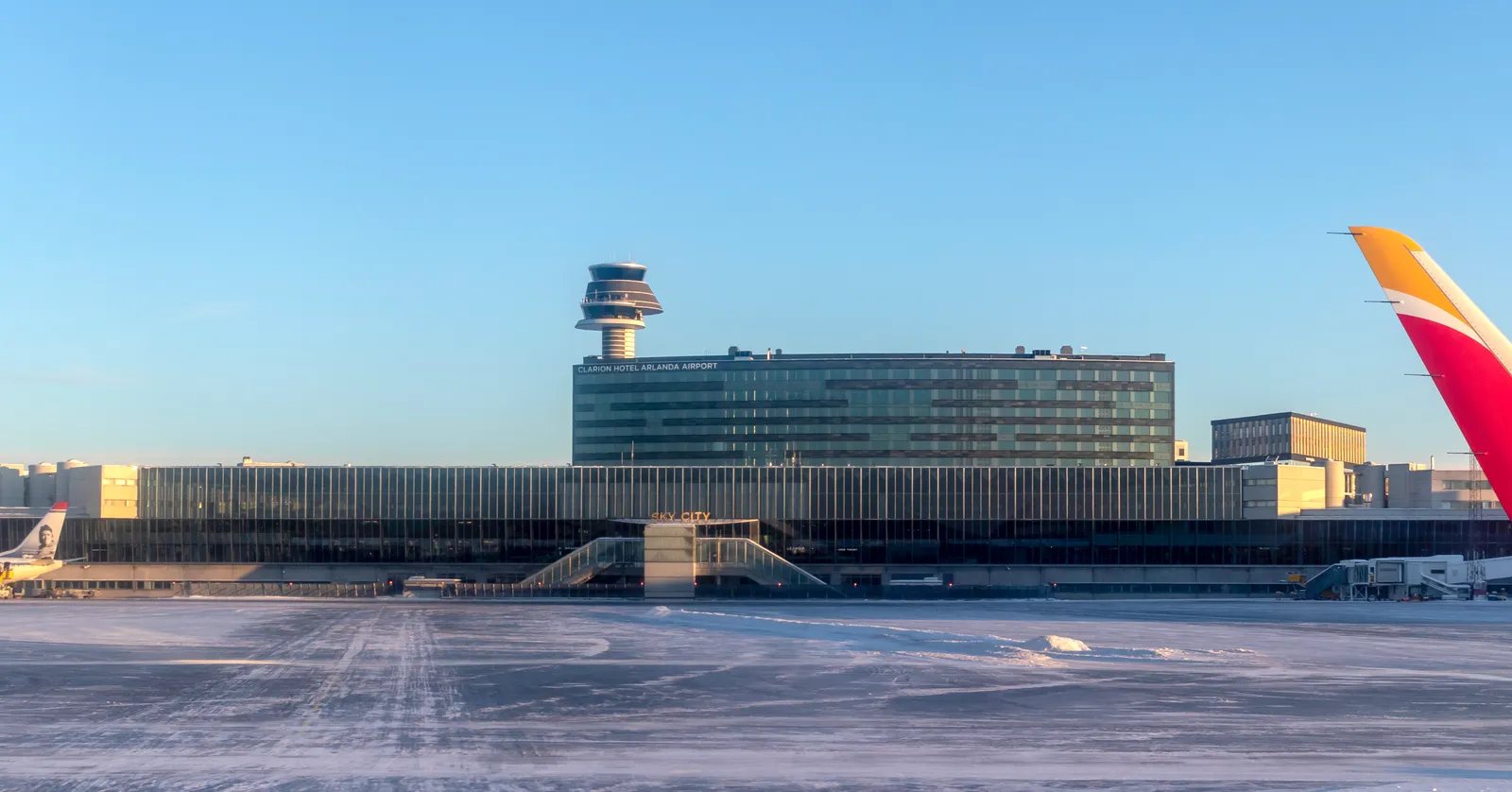 The Sky City block at Stockholm Arlanda Airport seen across a snow-edged apron, a long glass and steel building with the airport's slim control tower rising behind it under a clear winter sky