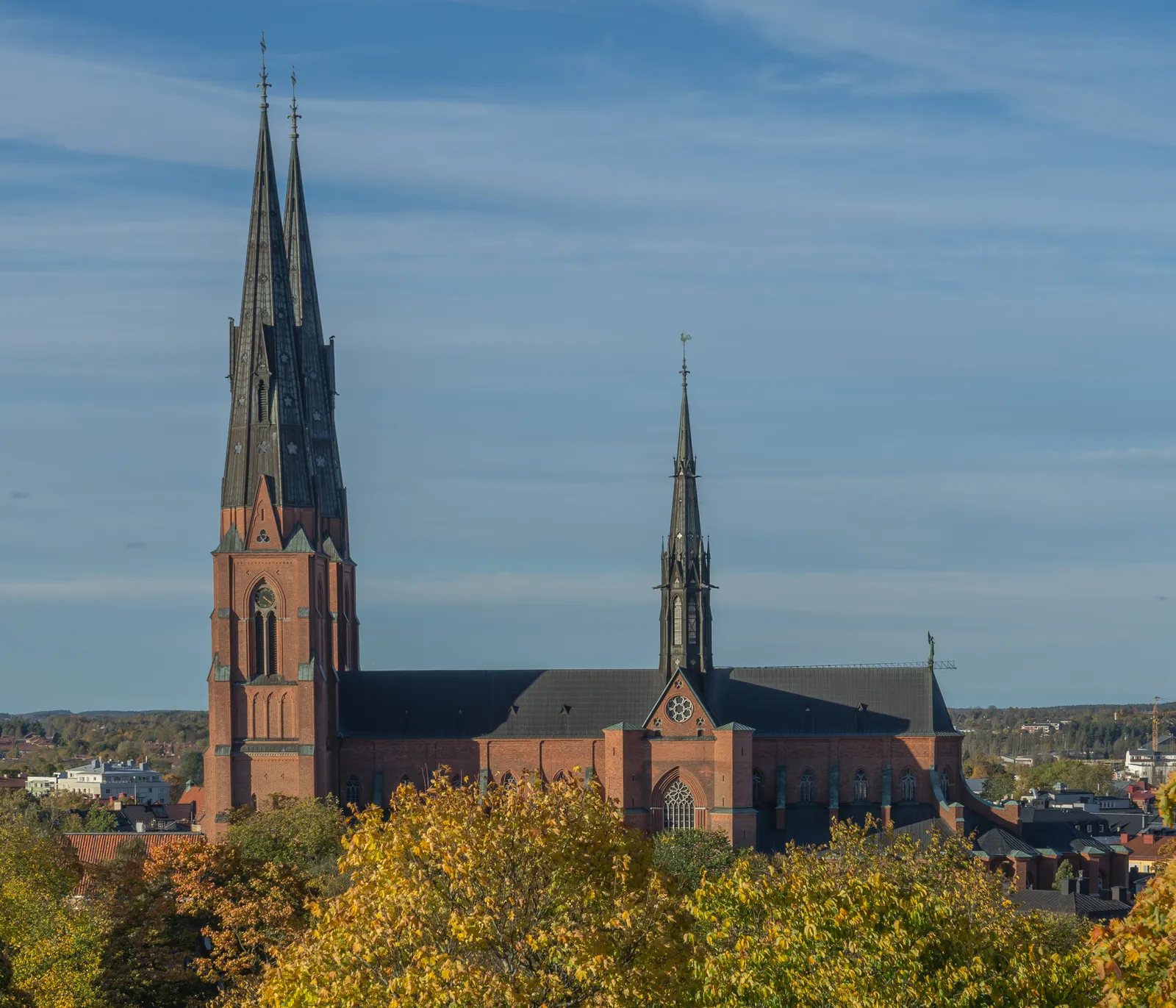 Uppsala Cathedral above autumn treetops, twin dark grey spires on a tall red brick tower front with the long nave and a smaller spire running back behind it