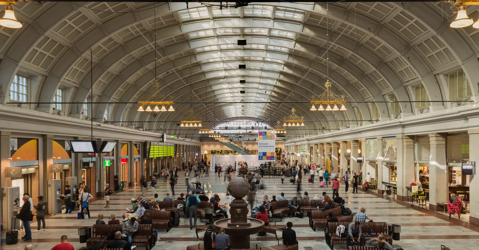 The main hall of Stockholm Central Station, a long barrel-vaulted room with arched glass roof panels, rows of hanging lamps and people sitting on benches around a stone fountain in the middle of the floor